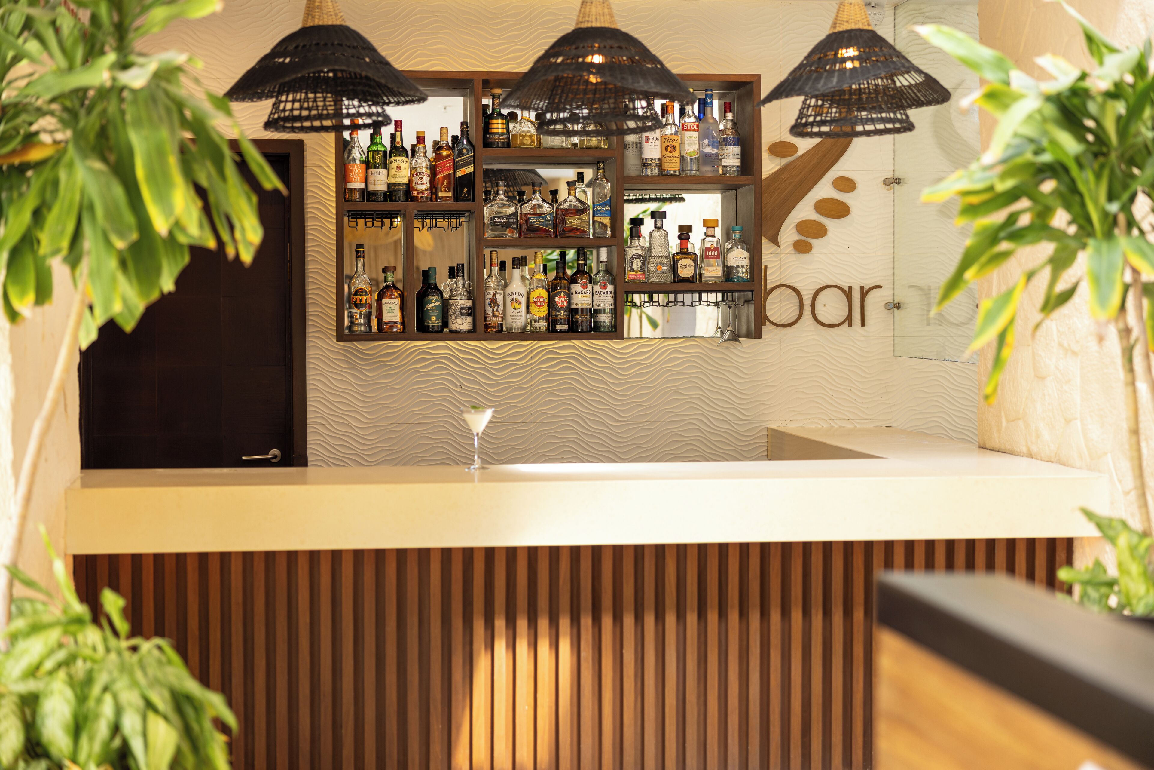 Zona de bar con estantes llenos de botellas de licores, una barra y lámparas colgantes.
