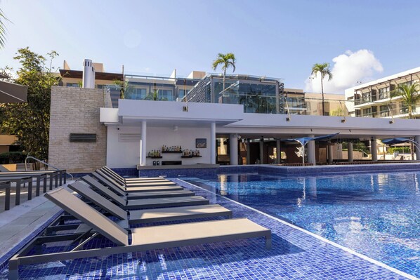A swimming pool with lounge chairs, a bar, and modern buildings in the background.