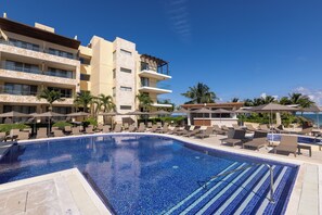 A swimming pool with lounge chairs and umbrellas, surrounded by multi-story buildings with balconies and palm trees. - Royalton Hideaway Riviera Cancun, An Autograph Collection All-Inclusive Resort- Adults Only - Newly Renovated (Puerto Morelos)