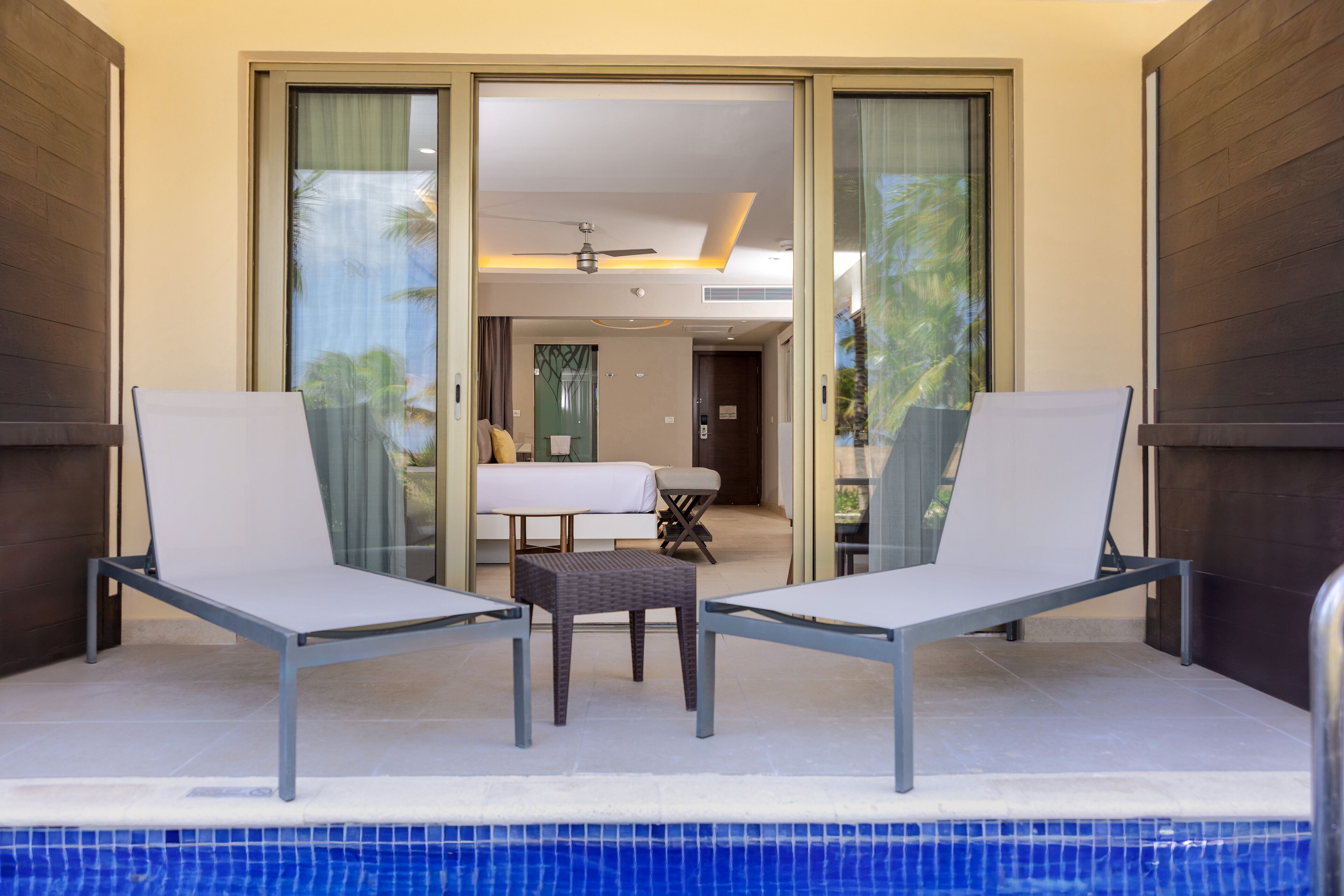 A pool area with two lounge chairs and a small table, a sliding glass door leading to a room, and a ceiling fan.