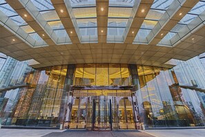 A modern building entrance with a glass facade, a central revolving door, and a geometric ceiling design.