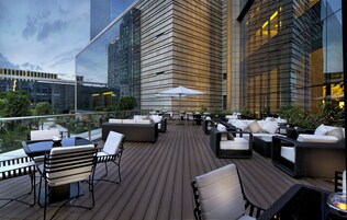 Terraza en la azotea con mobiliario exterior, barandilla de vidrio y vistas a un paisaje urbano.