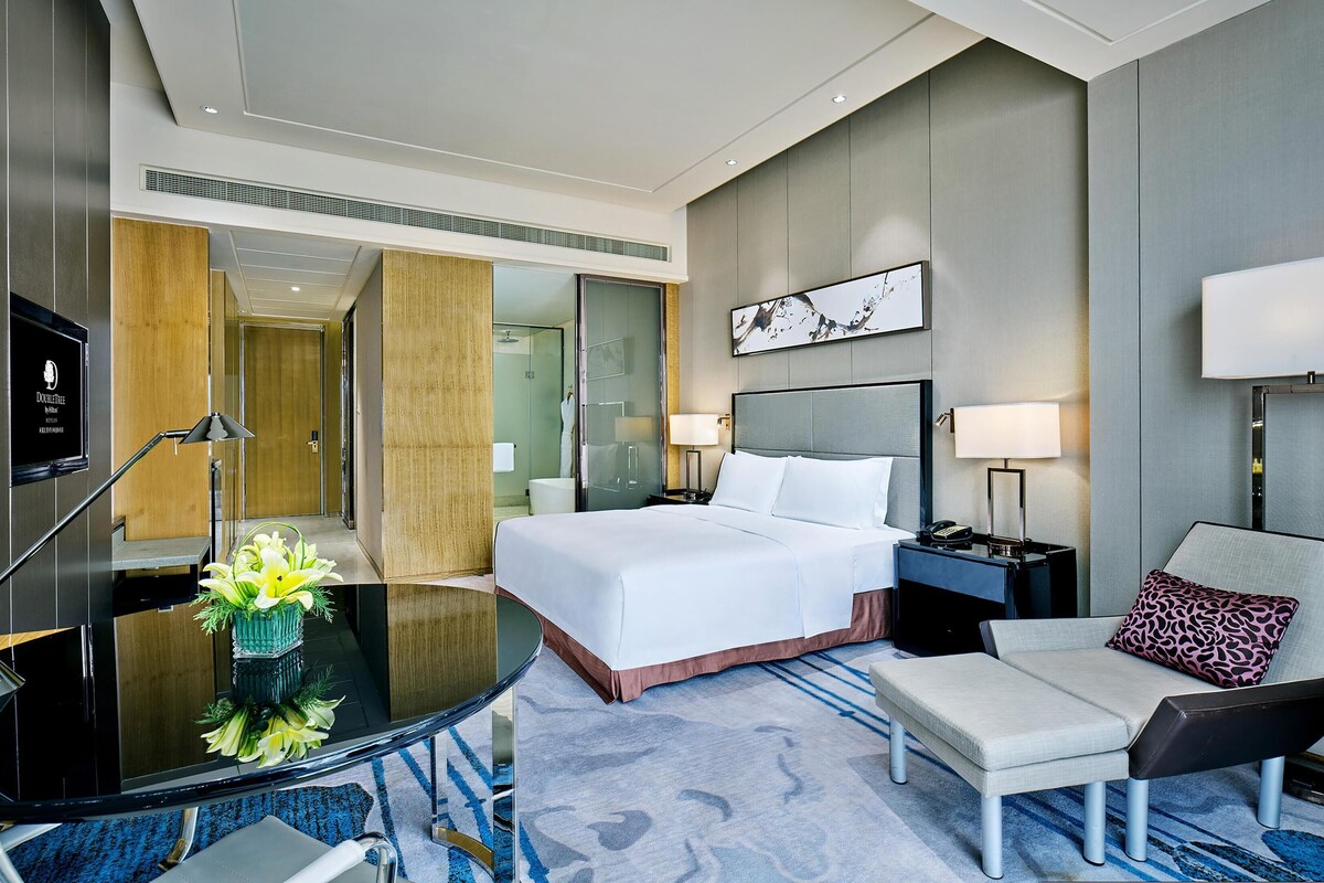A hotel room with a bed, bedside tables, a desk with a lamp, a chair, a glass shower enclosure, and a television.
