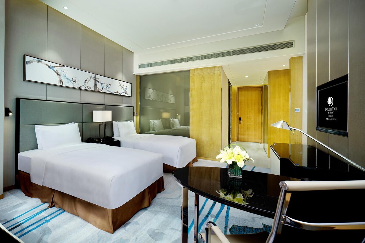 A hotel room with two beds, a desk with a chair, a TV, and artwork on the wall.