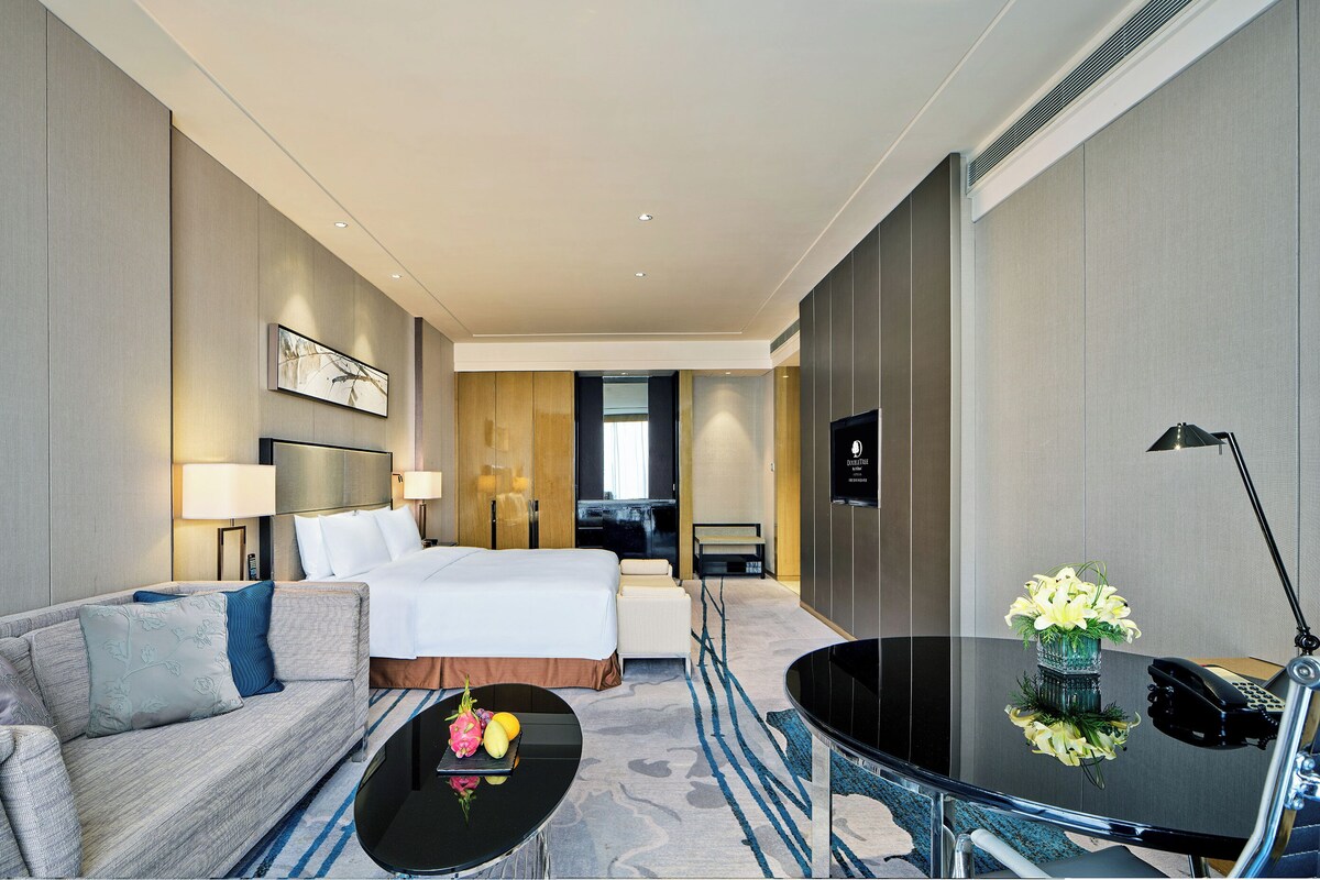 A hotel room with a bed, a sofa, a coffee table with fruit, a desk with a lamp, and a view of the hallway.