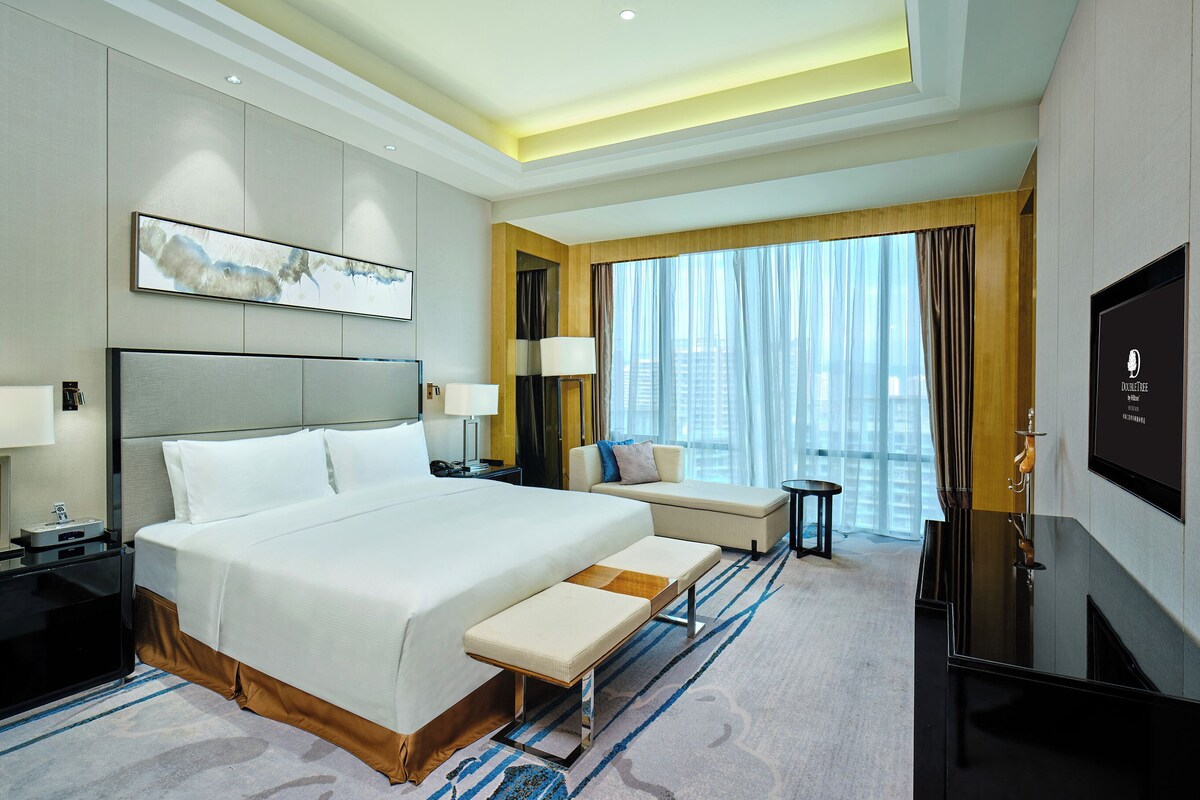 A modern hotel room with a large bed, a seating area, a television, and a city view.