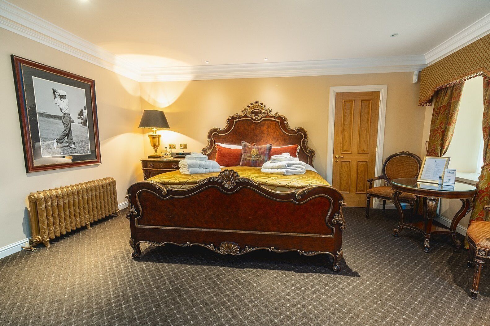 A traditional bedroom with a four-poster bed, a bedside table with a lamp, a framed picture on the wall, and a chair by the window.
