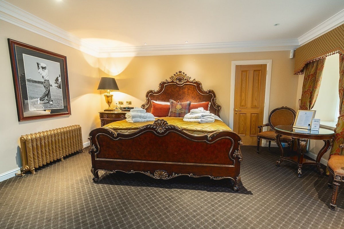 A traditional bedroom with a four-poster bed, a bedside table with a lamp, a framed picture on the wall, and a chair by the window.