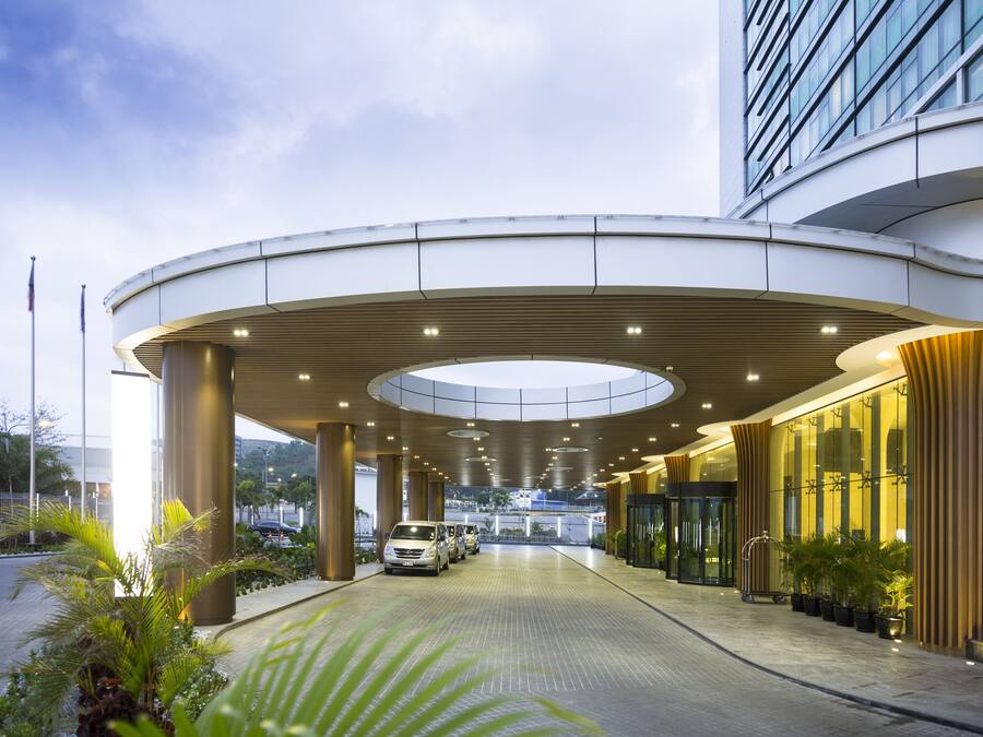 Ingresso moderno di un hotel con una tettoia curva, ampie colonne e un viale lastricato.