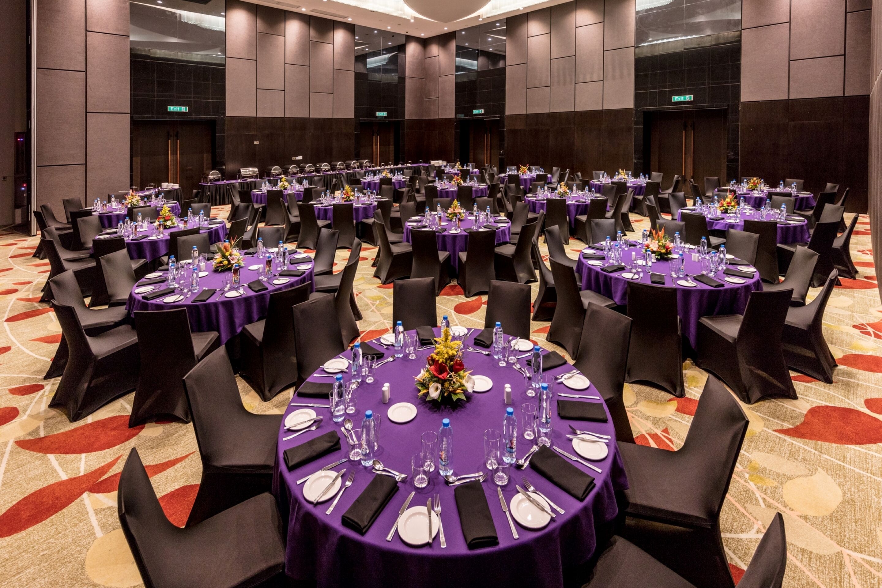 A large conference room with multiple round tables, each set with chairs, water bottles, and floral centerpieces.