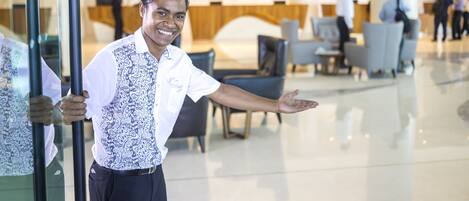 A man in a white shirt and patterned vest standing in a lobby with glass doors, a reception desk, and seating areas in the background.