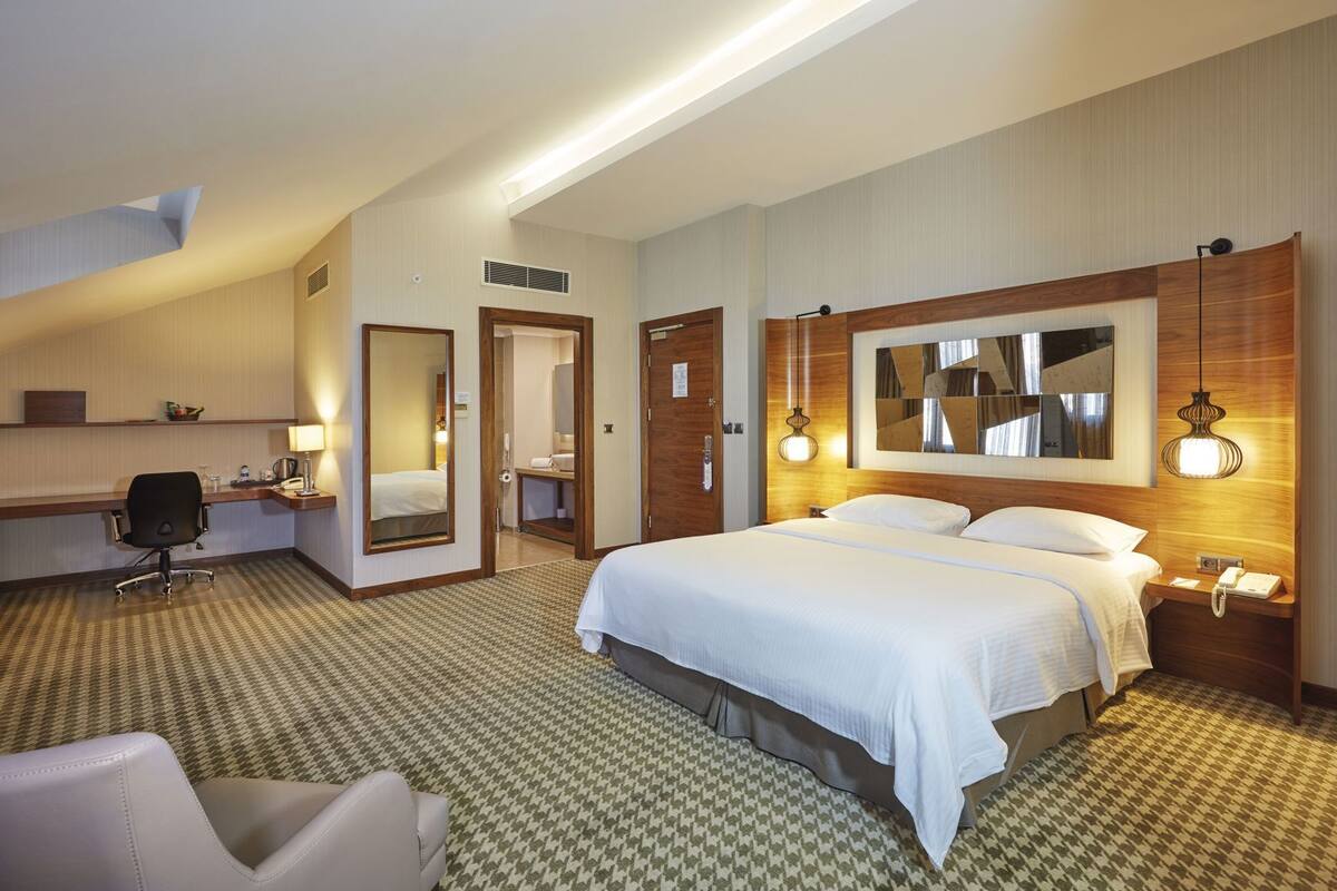 Una habitación de hotel con una cama grande, un escritorio, una silla, un espejo y una lámpara fijada a la pared.