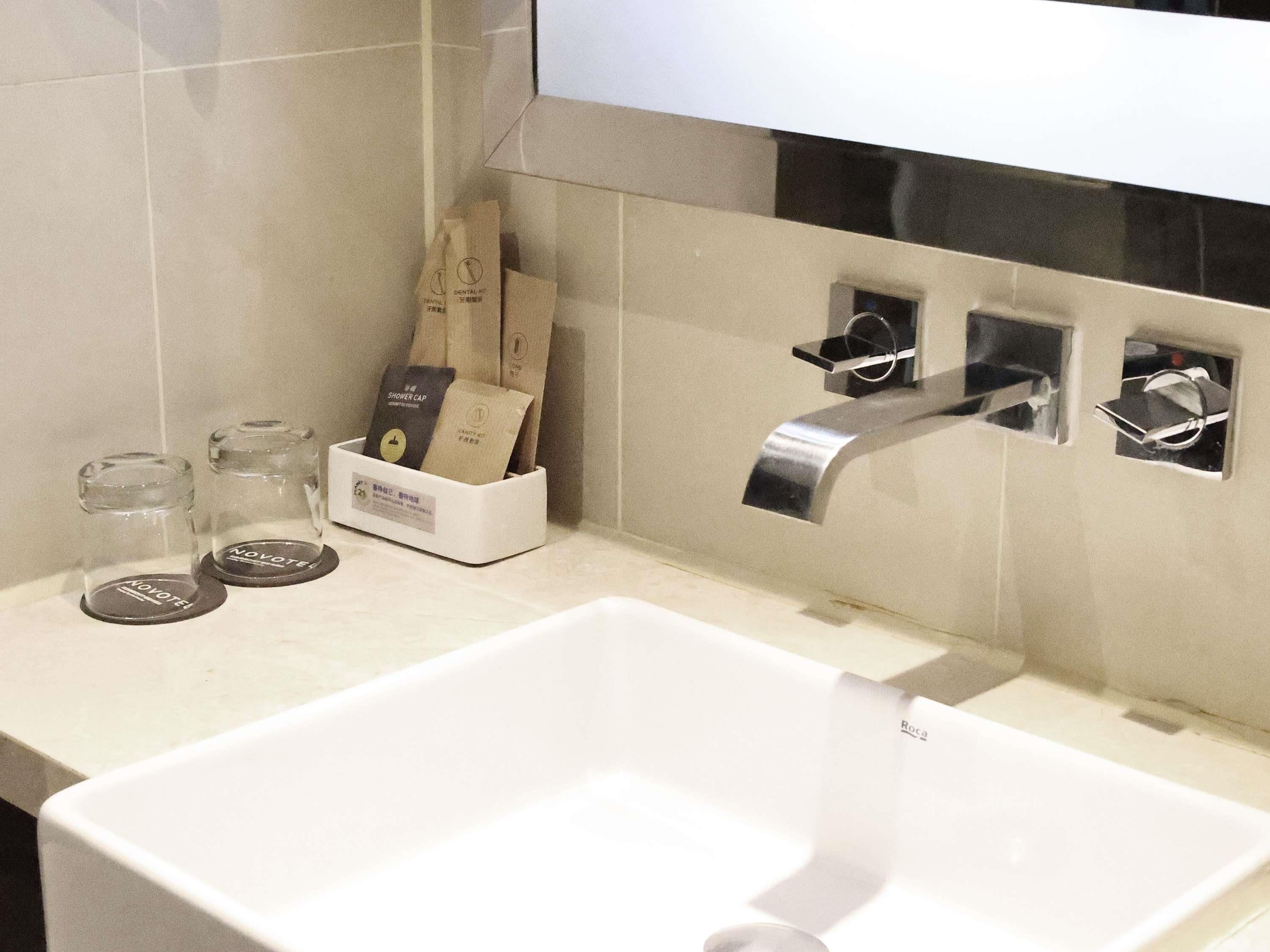 Un baño con un lavamanos blanco, un espejo y un perchero. Se ven dos botellas de vidrio y una caja blanca con etiqueta.