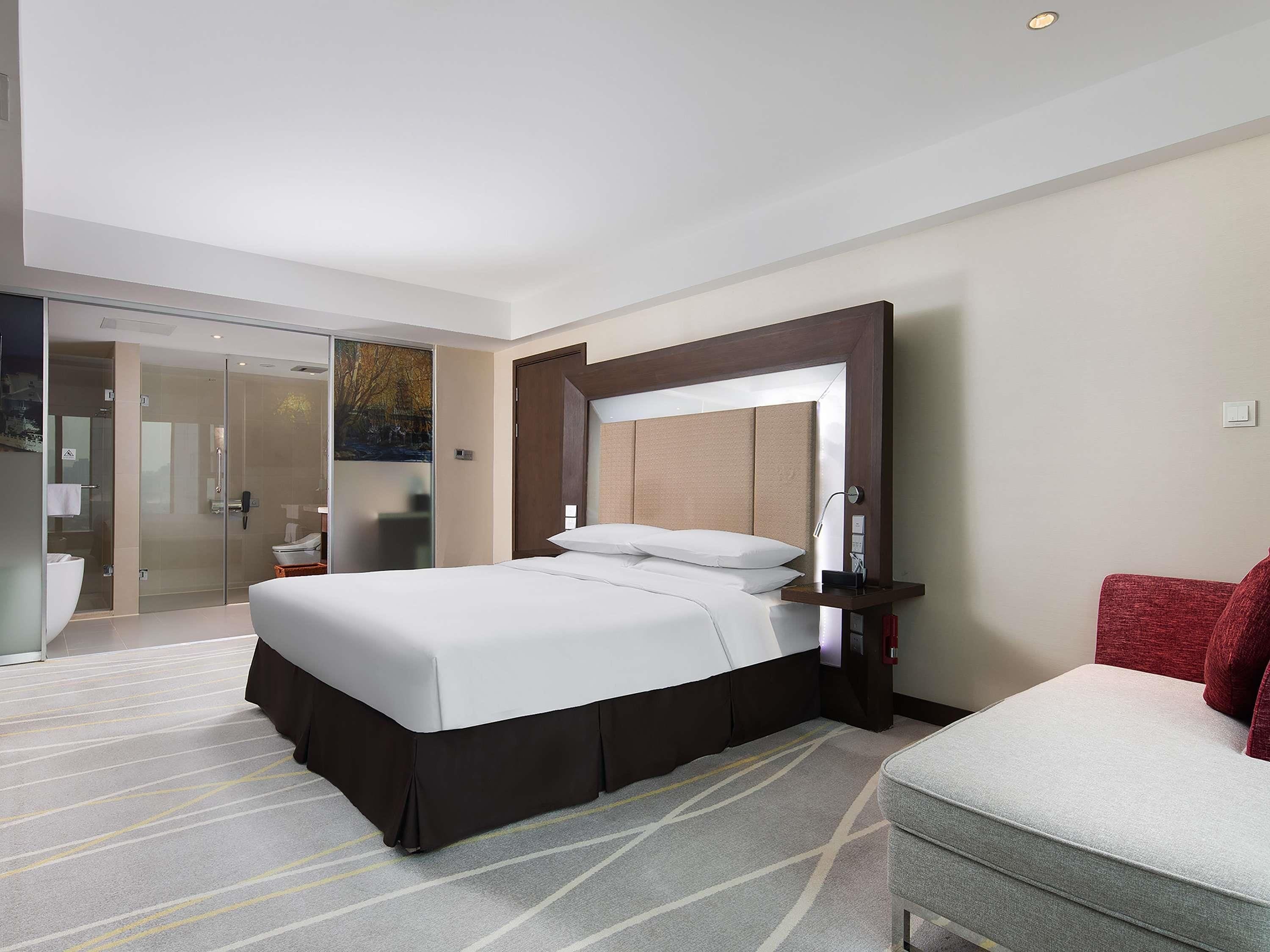 A modern hotel room with a large bed, a glass shower enclosure, a red armchair, and a large mirror above the bed.