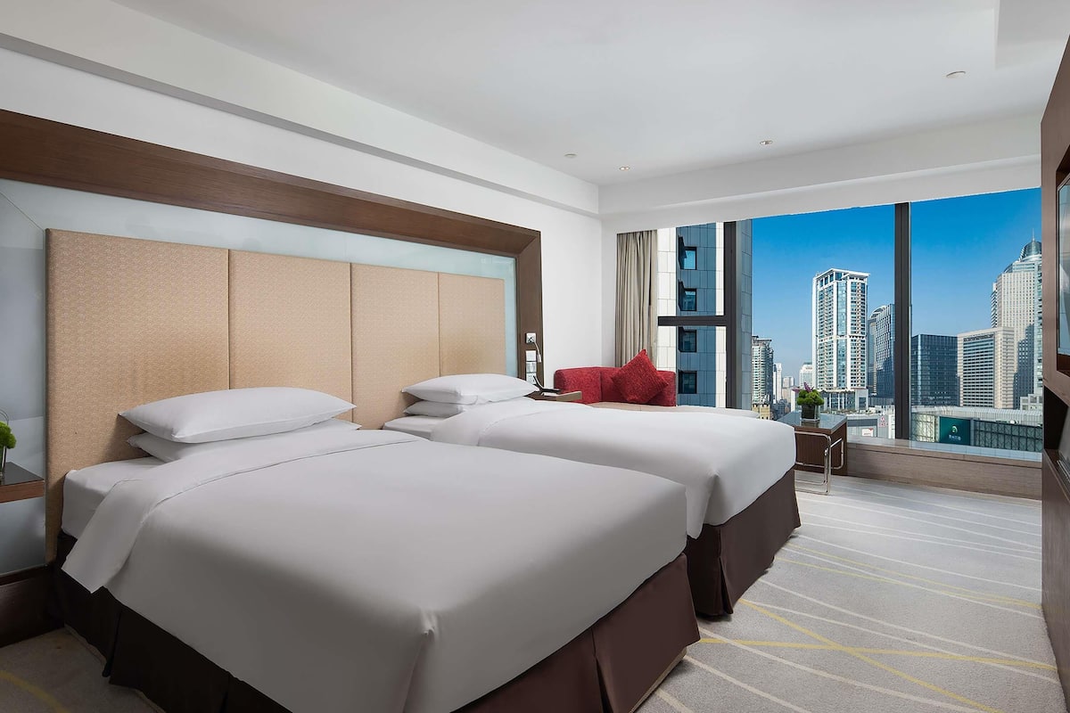 A hotel room with two beds, a large window with a city view, a red chair, and a small table with a vase.