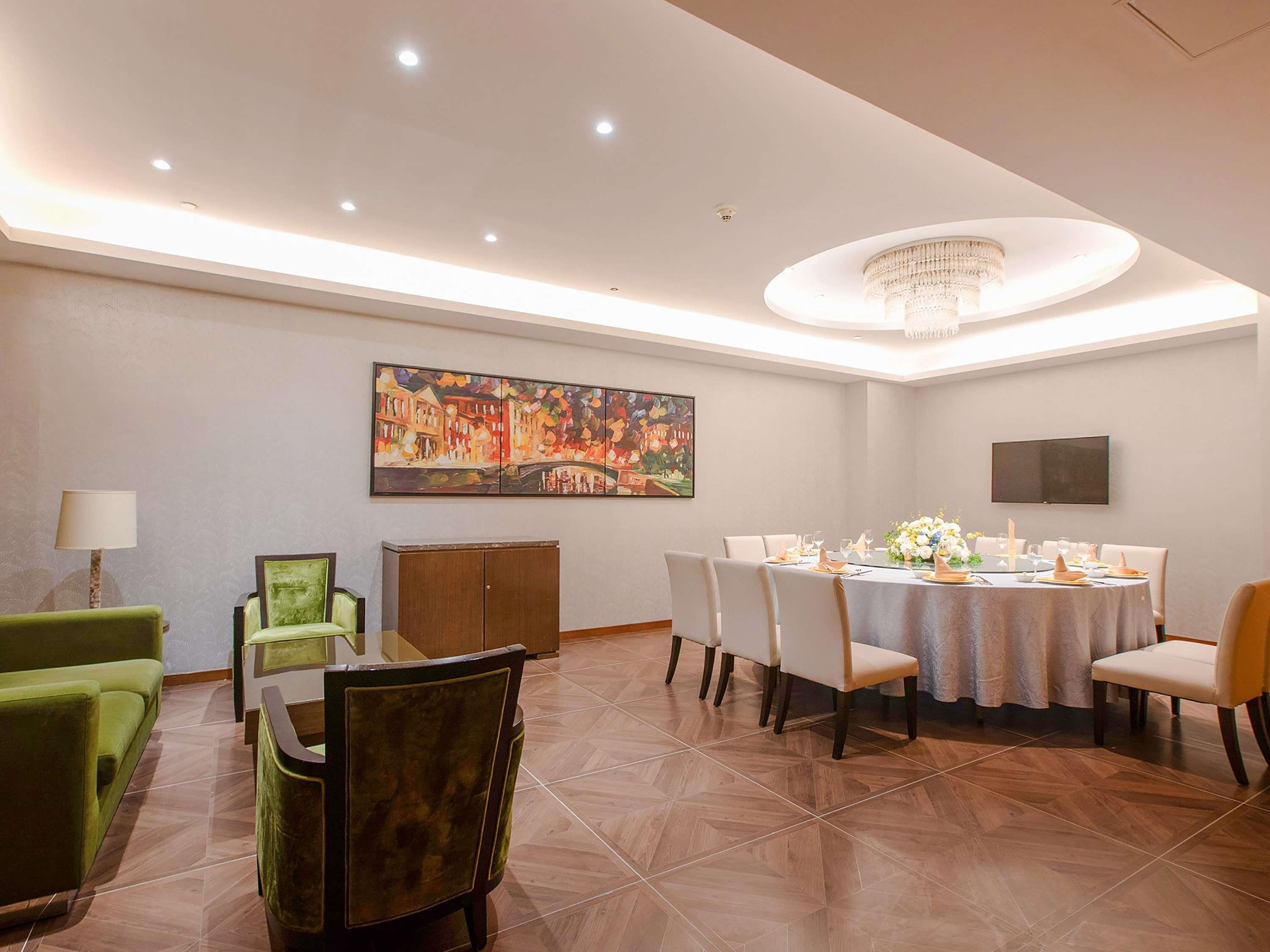 A dining area with a round table set for six, surrounded by chairs. There is a chandelier above the table, a painting on the wall, and a television mounted on the wall.
