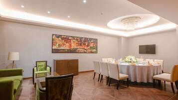 A dining area with a round table set for six, surrounded by chairs. There is a chandelier above the table, a painting on the wall, and a television mounted on the wall.