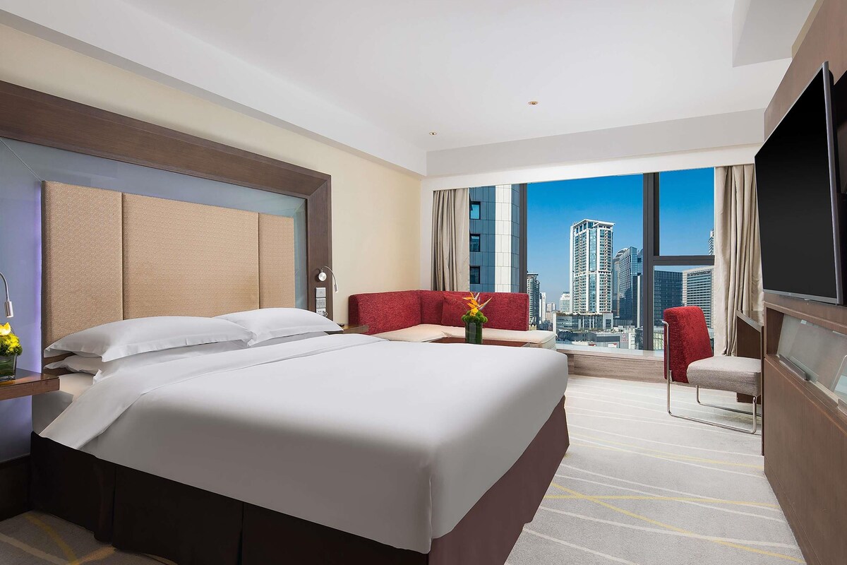 A hotel room with a large bed, a red sofa, a TV, and a city view.