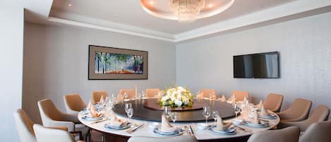 Een vergaderruimte met een ronde tafel, beige stoelen, een kroonluchter, een schilderij en een flatscreen televisie.