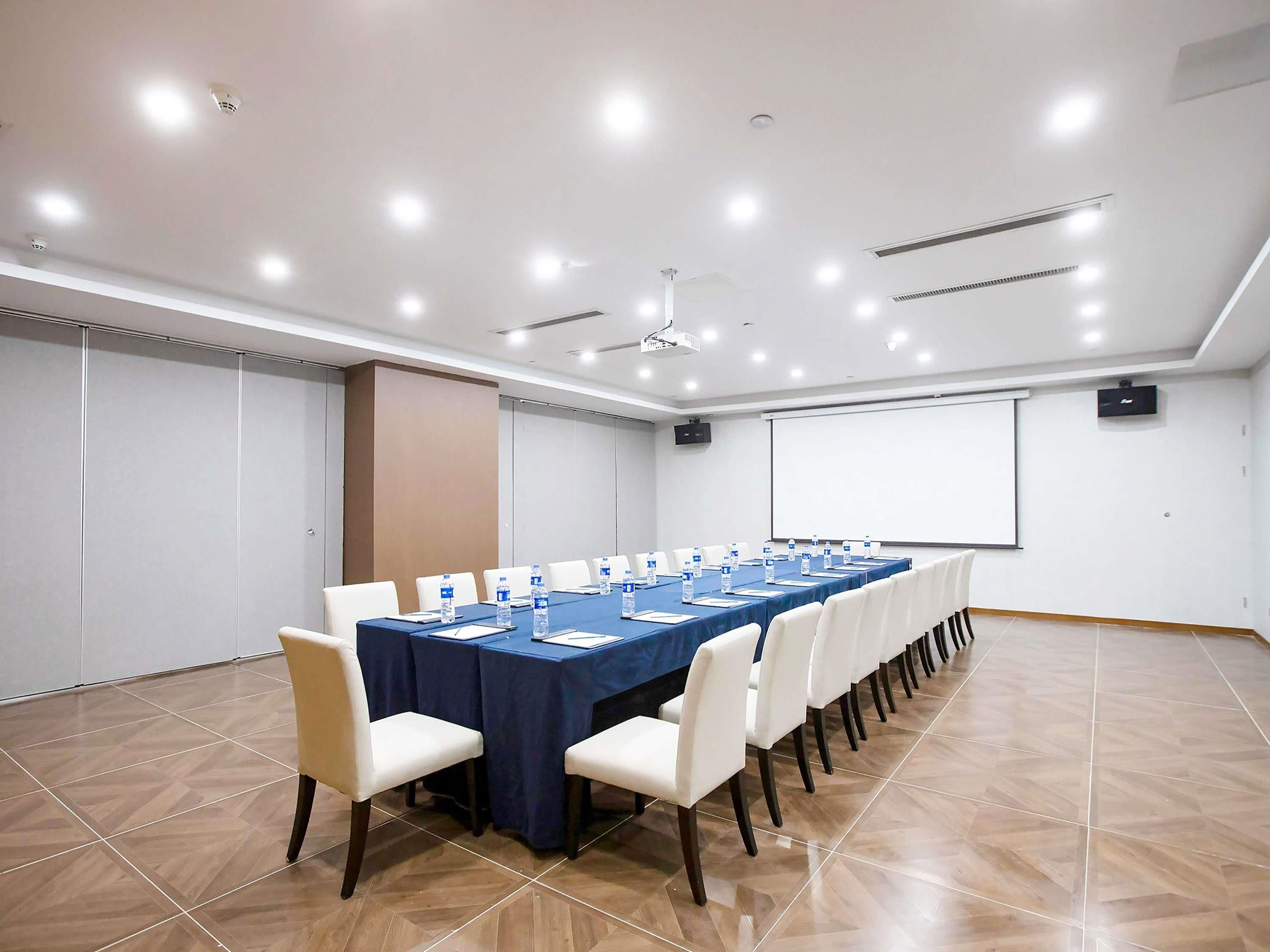 Un salón de conferencias con una larga mesa, sillas, una pantalla de proyector y una pizarra.