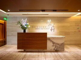 Un mostrador de recepción con panel de madera y un arreglo floral. La pared de fondo tiene un acabado texturizado y un letrero que dice 'ZIJIN GARDEN CHINESE RESTAURANT'.