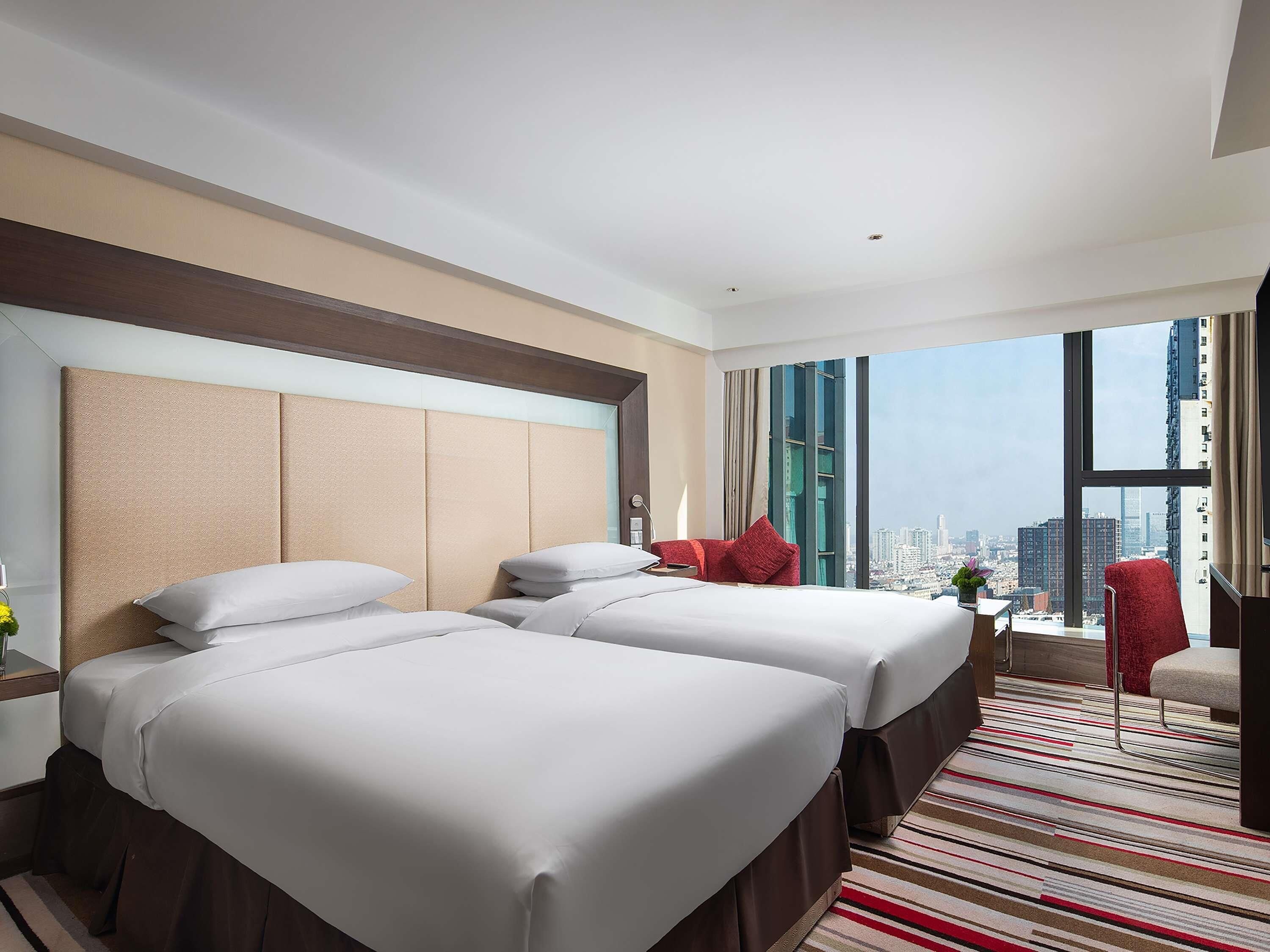 Une chambre d’hôtel avec deux lits, une grande fenêtre donnant sur la ville, un fauteuil rouge et une petite table avec une plante.