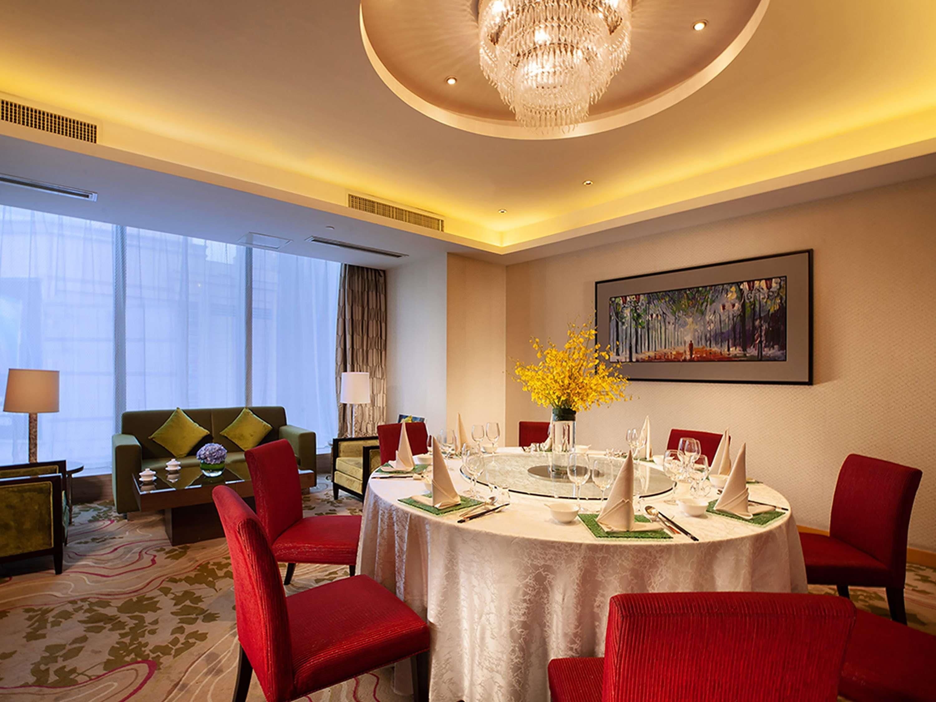 Un comedor de hotel con una mesa redonda preparada para seis comensales, rodeada de sillas rojas, un sofá con cojines verdes y un cuadro en la pared.