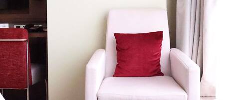 Un sillón blanco con un cojín rojo en una habitación con un televisor de pantalla plana montado en la pared y una silla roja al fondo.