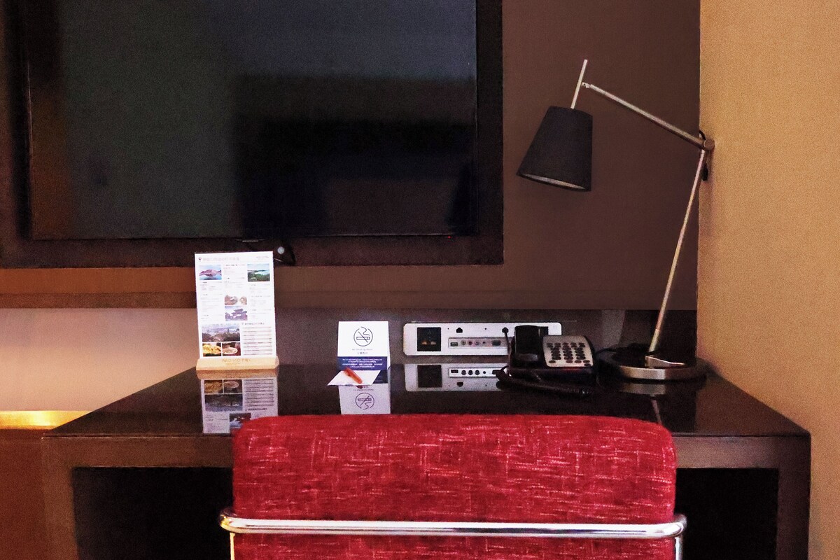 Habitación de hotel con televisor de pantalla plana montado en la pared, un escritorio con lámpara, teléfono y una silla roja.