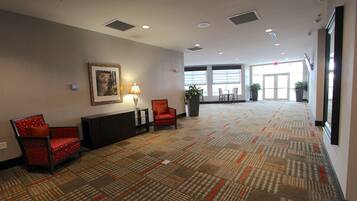 A spacious lobby with a patterned carpet, two red chairs, a desk with a lamp, a framed picture on the wall, and large windows allowing natural light.