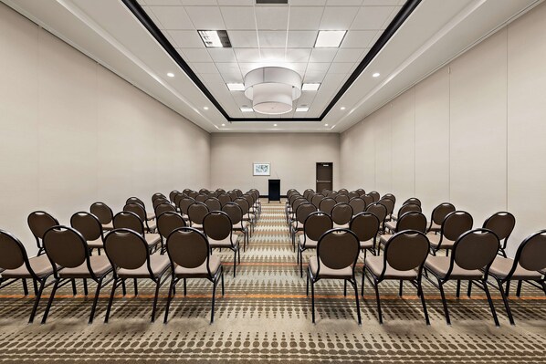 A conference room with rows of chairs facing a presentation screen, a carpeted floor, and a ceiling with recessed lighting.