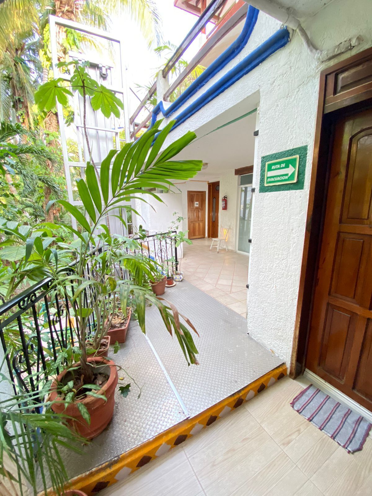 Entrada de un edificio con un letrero de salida de emergencia verde, plantas en macetas y una barandilla metálica.