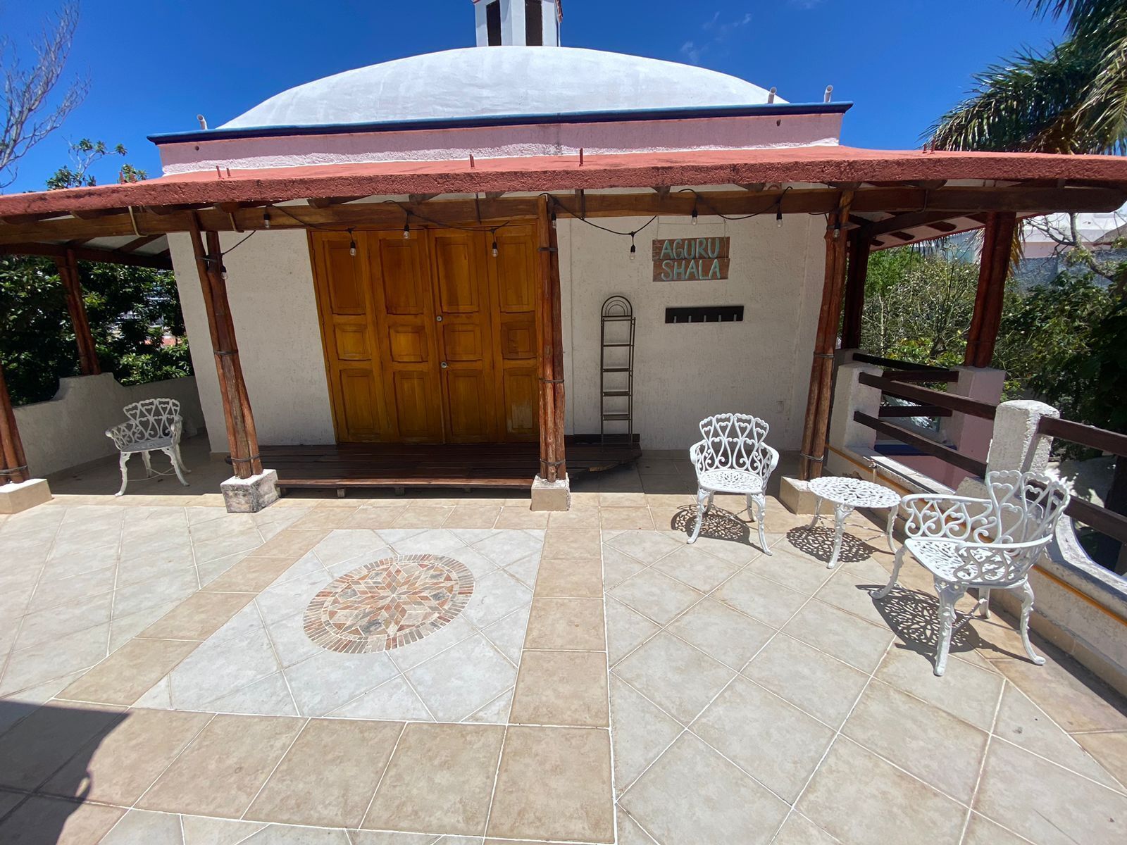 A covered patio with a tiled floor, a wooden door, and white metal chairs.