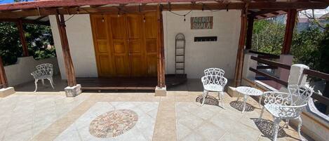Un patio techado con piso de baldosas, una puerta de madera y sillas de metal blanco.