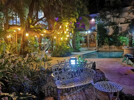 Un área de estar al aire libre con mobiliario de hierro forjado, rodeada de abundante vegetación y luces colgantes.