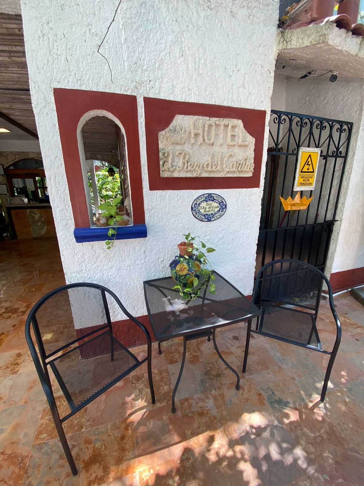 A hotel exterior with a sign reading "HOTEL El Rey de Caritas," a small table with chairs, and a potted plant.