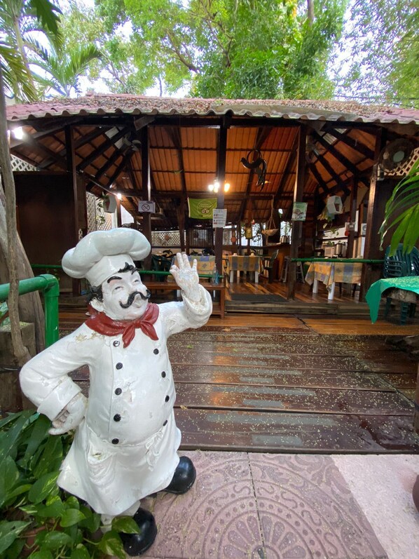 A rustic outdoor seating area with a wooden structure, a statue of a chef, and a patterned floor.