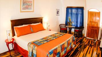 A hotel room with a wooden floor, a bed with orange bedding, a wooden headboard, a small table with chairs, and a door.