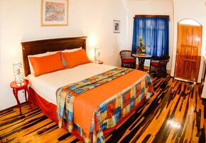 A hotel room with a wooden floor, a bed with orange bedding, a wooden headboard, a small table with chairs, and a door.