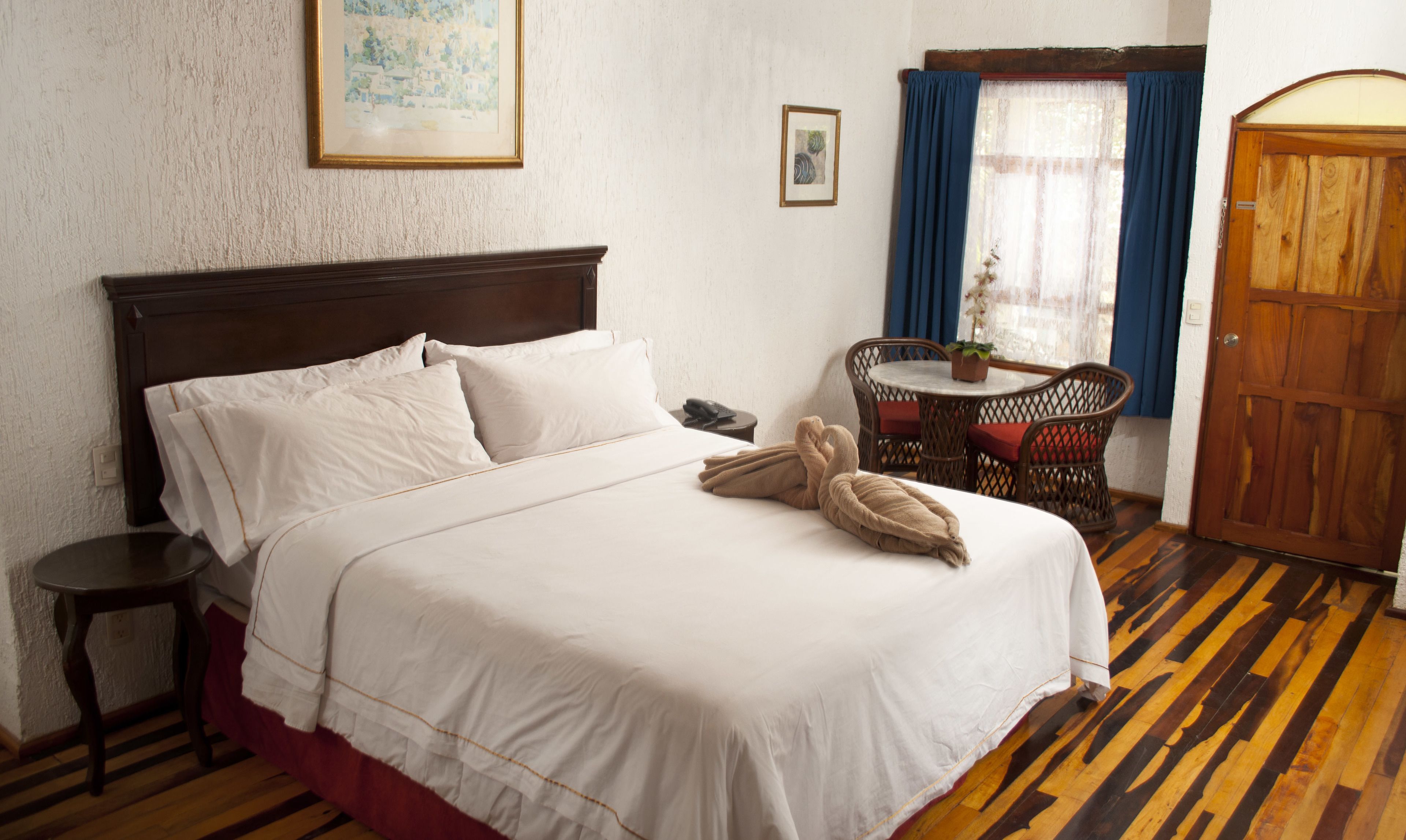 Uma cama bem arrumada com lençóis brancos, cabeceira de madeira, pequena mesa de cabeceira e uma cadeira de vime com mesa.