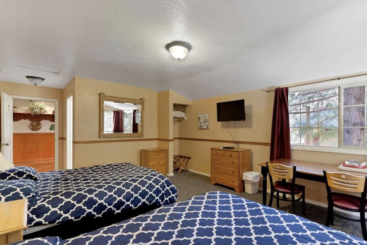 Quarto de hotel com duas camas, uma escrivaninha, uma cadeira, televisão e janela vista para árvores.
