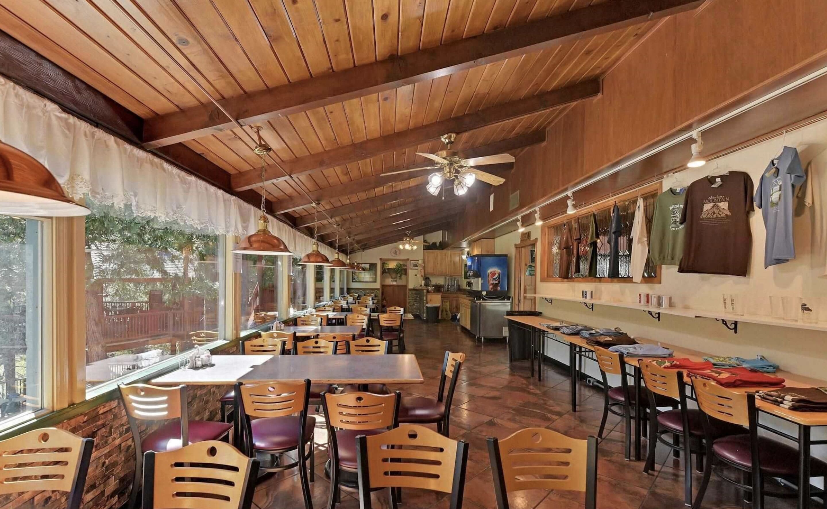 Um restaurante com vigas de madeira no teto, janelas amplas e uma área de cozinha visível ao fundo.