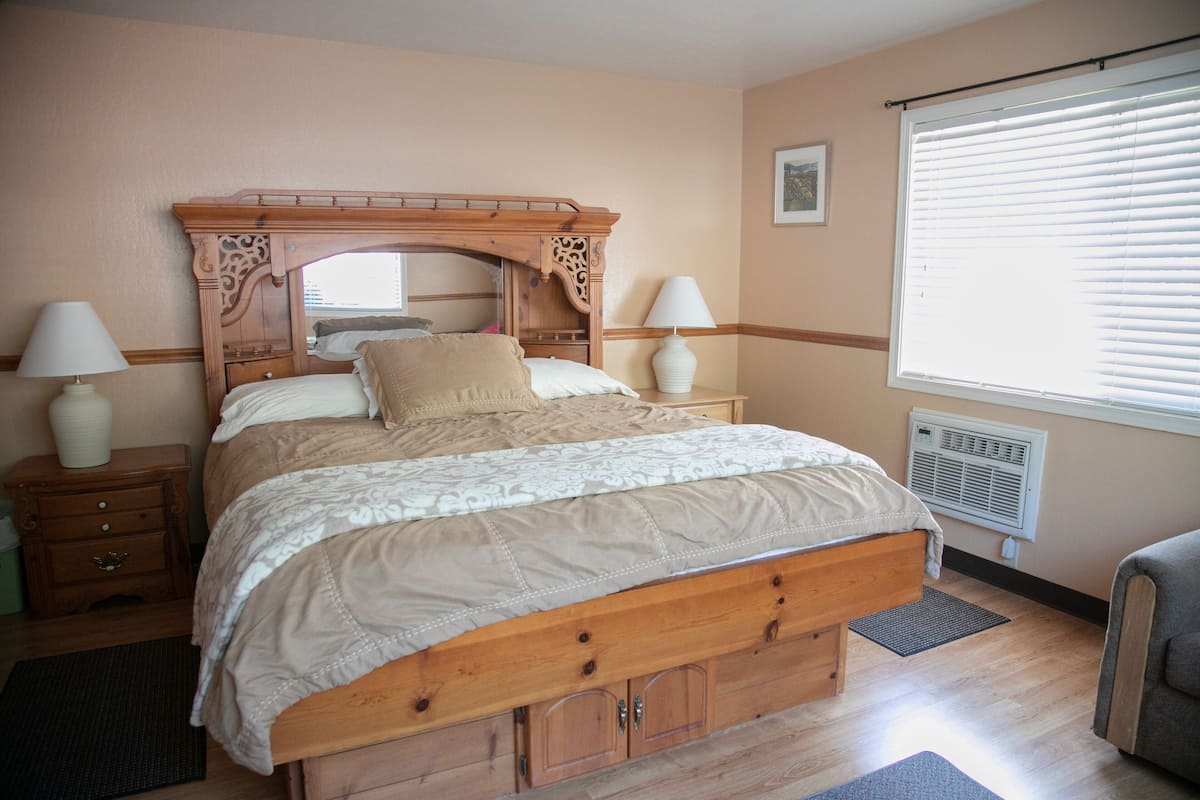 Quarto com cama de madeira, duas mesas de cabeceira com luminárias, janela com persianas e ar-condicionado fixado na parede.