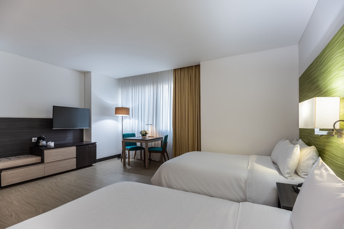 A hotel room with two beds, a desk with a chair, a television, and a lamp.