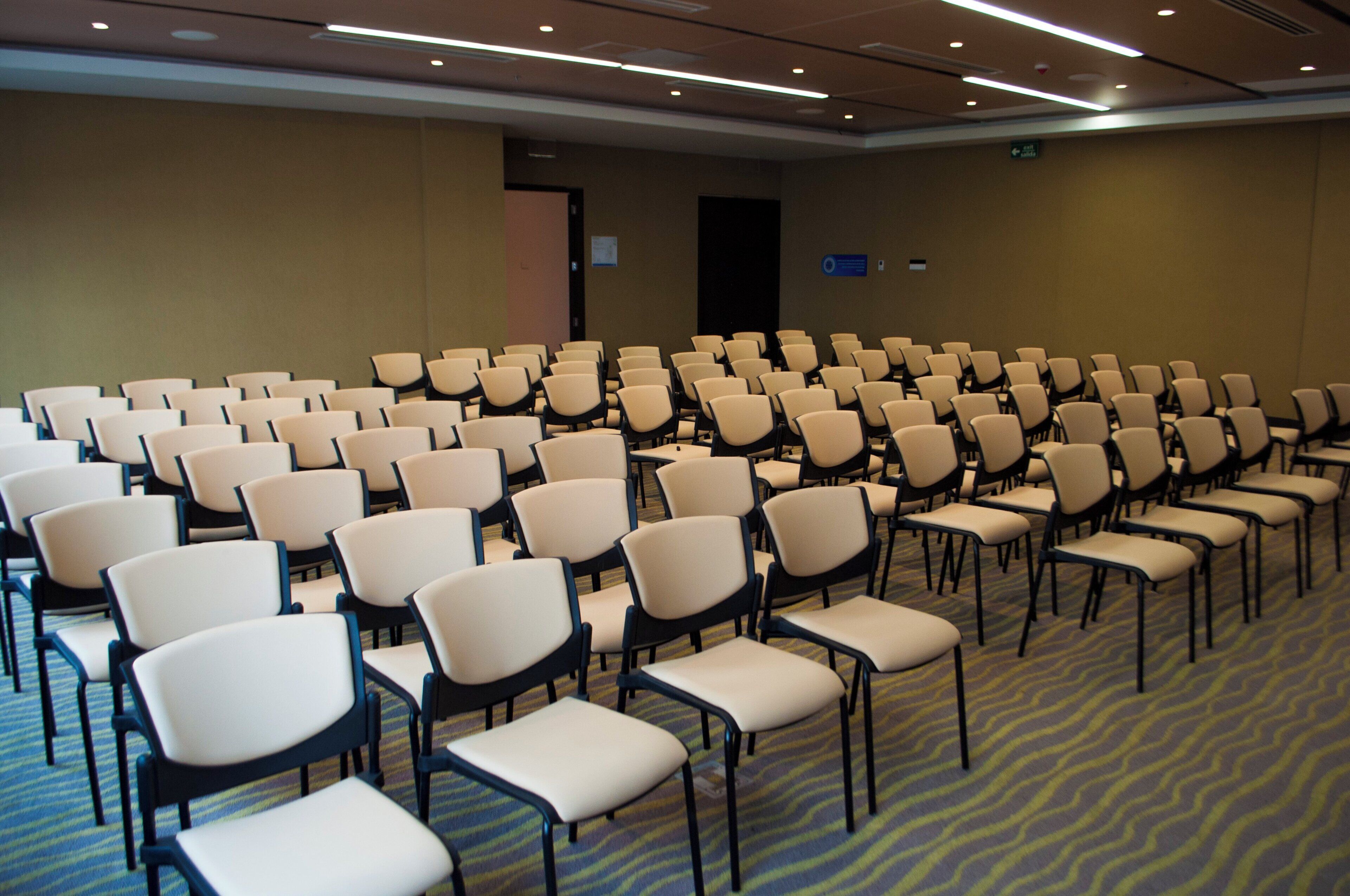 Une salle de conférence avec des rangées de chaises, un sol recouvert de moquette et une porte au fond.