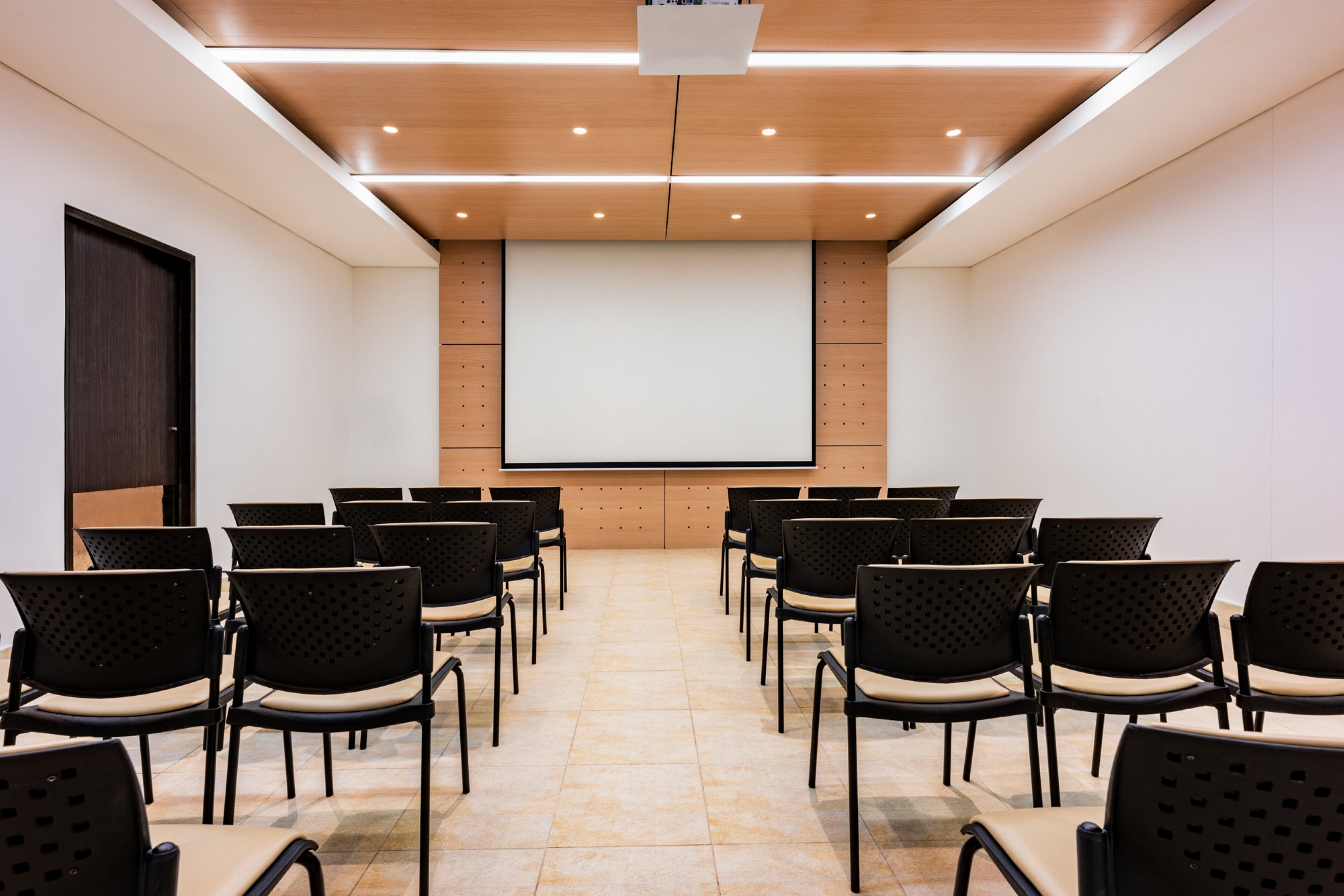 Un salón de conferencias con filas de sillas orientadas hacia una pantalla grande, paredes de paneles de madera e iluminación empotrada en el techo.