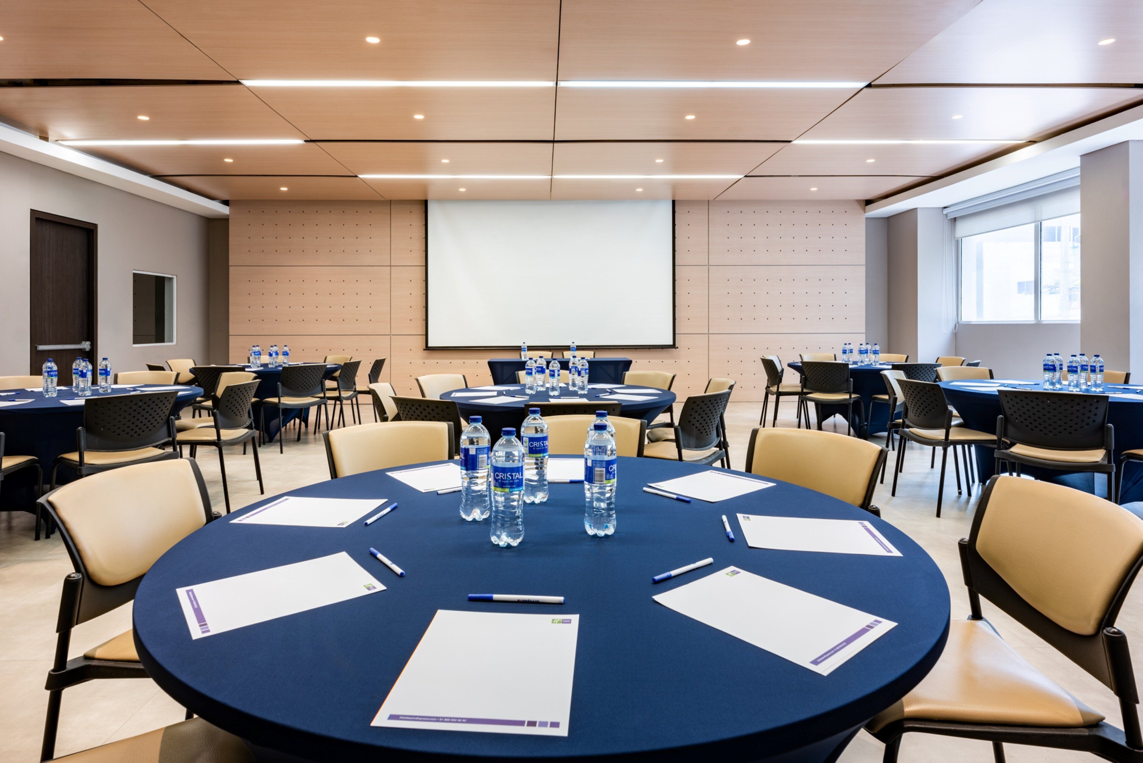 Une salle de conférence équipée de tables rondes, de chaises, de bouteilles d’eau et d’écrans de présentation.