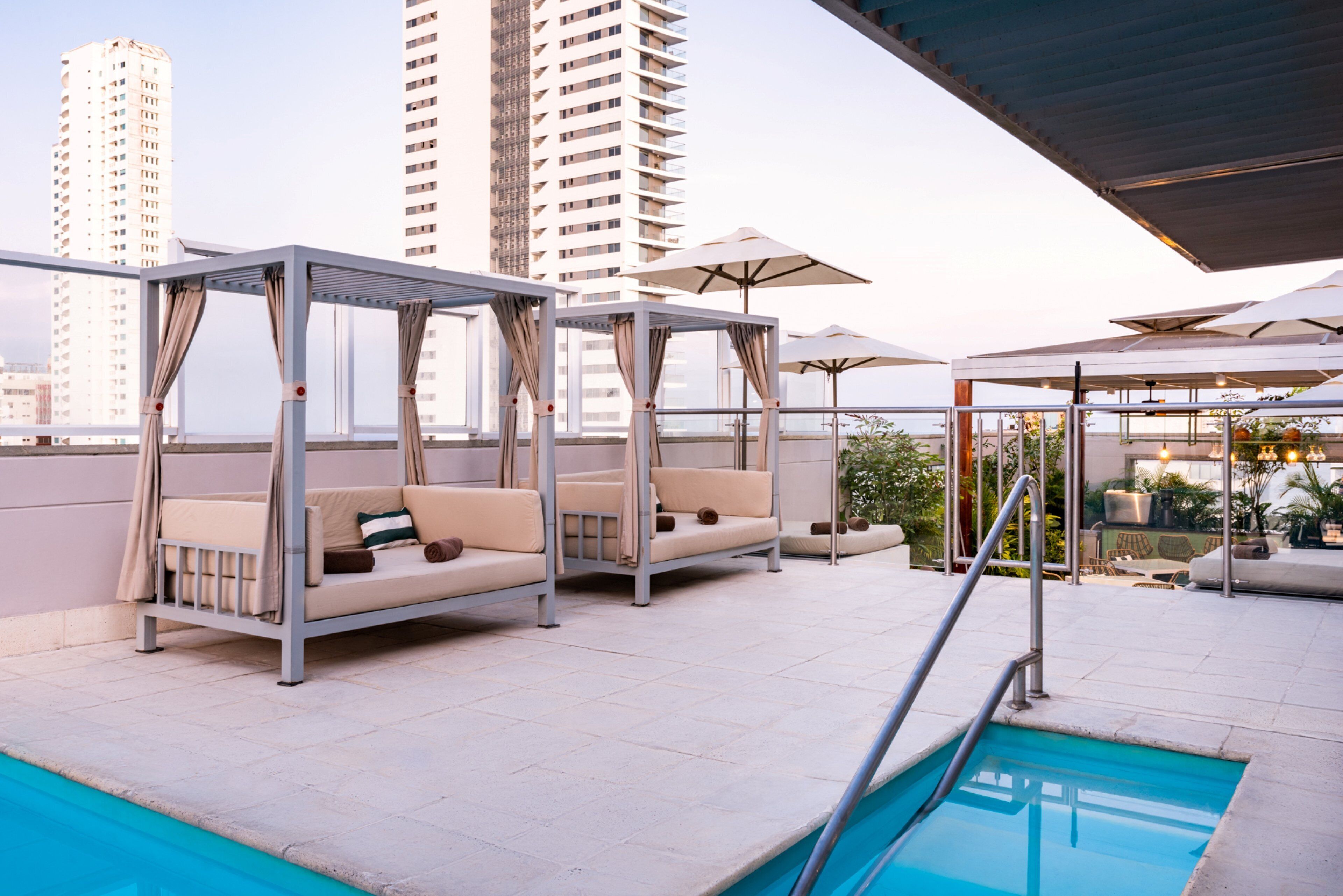 A rooftop pool area with outdoor seating and umbrellas, overlooking a cityscape.