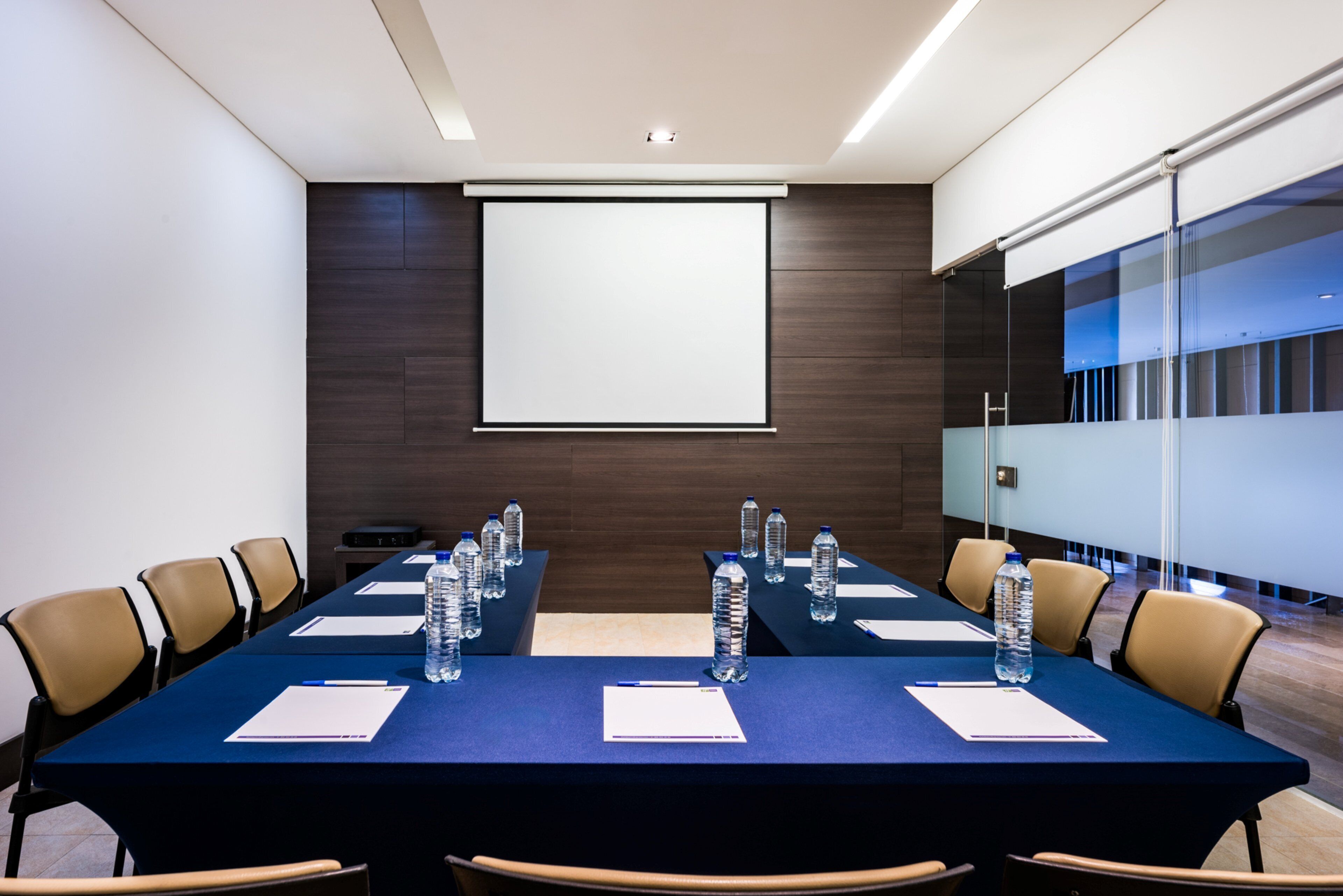 A conference room with a long table, chairs, water bottles, and a projector screen.