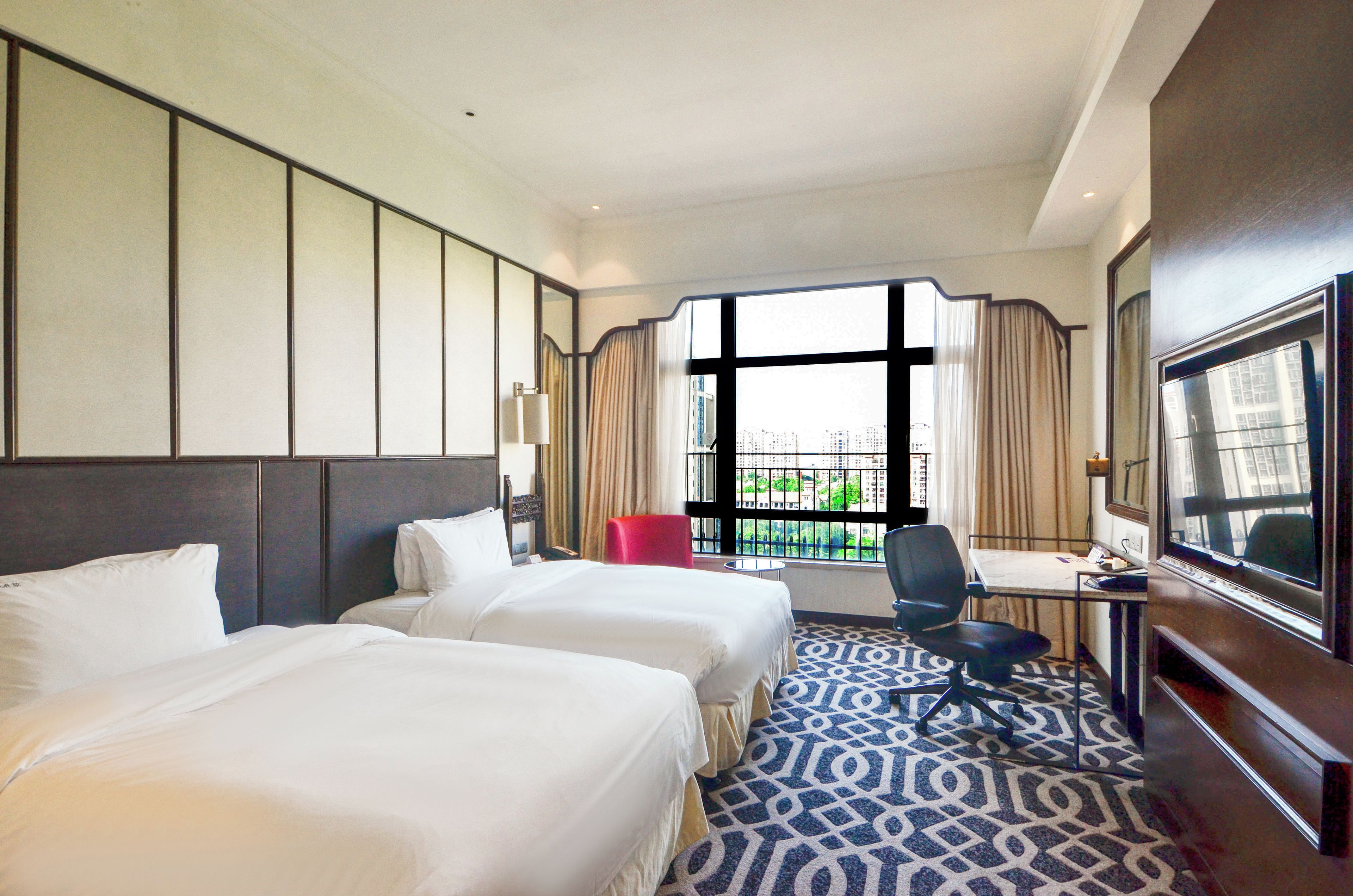 A hotel room with two beds, a desk, a chair, a TV, and a large window with a city view.